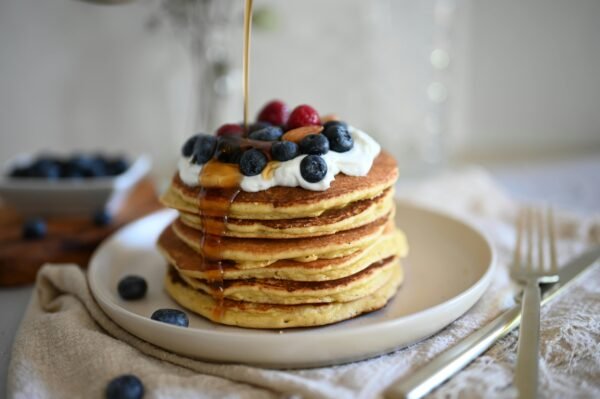 Stack of pancakes with blueberries and raspberries topped with syrup and whipped cream on a plate.